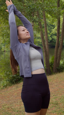 Vertical-Video-Of-Woman-Wearing-Sports-Clothing-Doing-Yoga-Stretches-On-Mat-In-Forest-By-Lake-Or-River-Enjoying-Peace-And-Beauty-Of-Nature-Shot-In-Real-Time